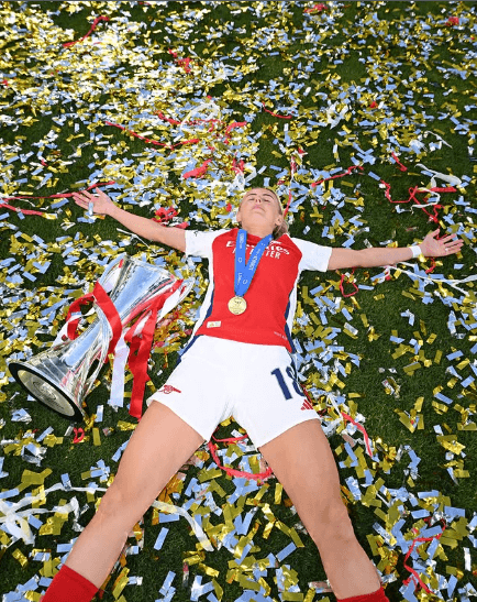 England women's football player lying on the field with a medal and trophy, celebrating back-to-back championship victory amid confetti